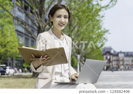 一位年輕的女商人坐在長椅上，一邊看著筆記型電腦，一邊在筆記本上寫字 128882406