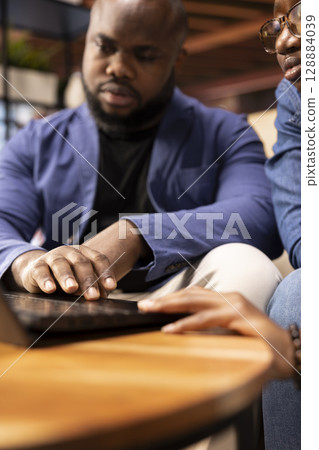 Casual man and woman opening laptop on the coffee table to solve daily tasks, doing remote freelance work online. Black couple surfing the web and multitasking in their loft apartment. Close up. 128884039
