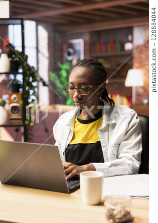 Student at home browsing internet on notebook, doing homework, researching information online. African american girl in apartment reading articles on laptop for school tasks Student at home browsing internet on notebook, doing homework, researching information online. African american girl in apartment reading articles on laptop for school tasks 128884044