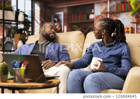 African American couple enjoys a break together and sipping coffee, engaging in chill conversation in their cozy living room. Their home lifestyle reflects love and quality time, couple goals. 128884045