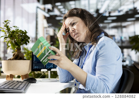 Businesswoman with a headache preparing to take a painkiller medicine in order to solve her daily organizational tasks, reading the recommended dosage on the box label. Aspirin pills. Businesswoman with a headache preparing to take a painkiller medicine in order to solve her daily organizational tasks, reading the recommended dosage on the box label. Aspirin pills. 128884082
