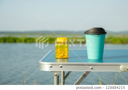 Yellow vitamin container and teal travel mug on blue camping table outdoors by the lake. 128884215
