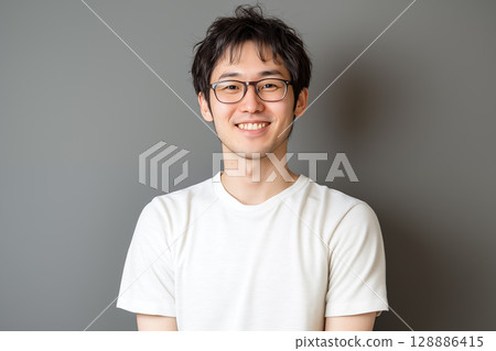 Portrait of a young Japanese man in a white T-shirt smiling in front of a gray background 128886415