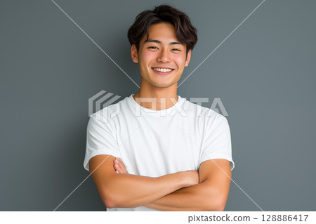 Portrait of a young Japanese man in a white T-shirt smiling in front of a gray background 128886417