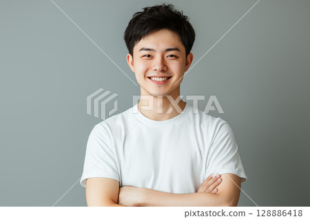 Portrait of a young Japanese man in a white T-shirt smiling in front of a gray background 128886418