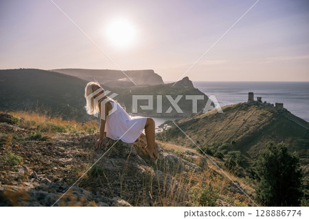 A woman is sitting on a hillside overlooking the ocean. She is wearing a white dress and has blonde hair. The scene is serene and peaceful, with the ocean in the background. 128886774