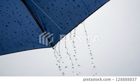 Blue umbrella with raindrops dripping from edge against overcast white sky during rainfall weather protection concept 128888837
