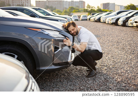 A man is happy, embracing his new car at a car dealership. He is kneeling and touching the front of the vehicle with affection. A man is happy, embracing his new car at a car dealership. He is kneeling and touching the front of the vehicle with affection. 128890595