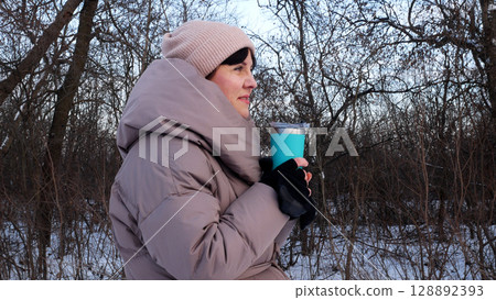 A young woman in a warm down jacket drinks tea or coffee from a thermos  128892393