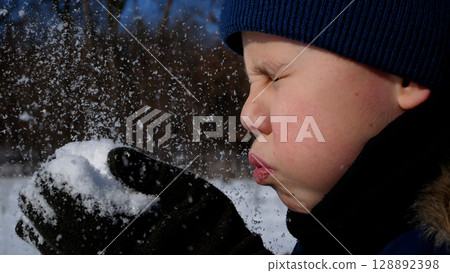 A boy blows on white snow in his hands in winter. The snow is scattering.  128892398