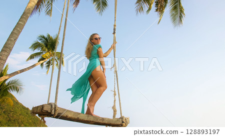 Seductive woman swinging on a swing on a tropical beach, on shores of the turquoise sea. Concept travel, walks, rest in sea, tropical resort coastline relaxation traveling tourism summer holidays. 128893197