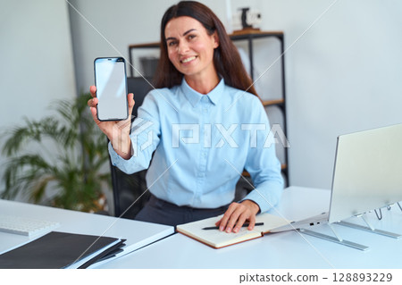 Professional Businesswoman Showing Smartphone Screen During Office Work in Modern Workspace Professional Businesswoman Showing Smartphone Screen During Office Work in Modern Workspace 128893229