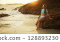 Woman leaning on a rock reef enjoying and relax warm dark golden sunset. Palm tree in beach. Woman in blue swimsuit and sunglasses Woman leaning on a rock reef enjoying and relax warm dark golden sunset. Palm tree in beach. Woman in blue swimsuit and sunglasses 128893230