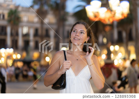 In evening, young smiling girl walks along city street Placa Real In evening, young smiling girl walks along city street Placa Real 128893308