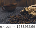 Burlap sack spilling coffee beans with blurred coffee cup background 128893369
