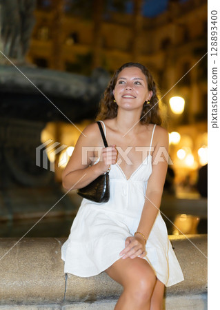 Night Royal square in Barcelona, girl in sundress is sitting on fountain fencing. Night Royal square in Barcelona, girl in sundress is sitting on fountain fencing. 128893400