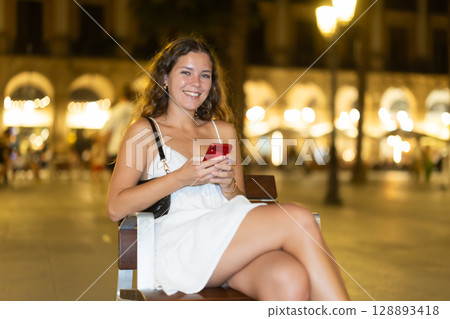 Night Royal square in Barcelona, girl in sundress with phone is sitting chair Night Royal square in Barcelona, girl in sundress with phone is sitting chair 128893418