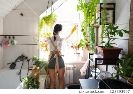 Woman in rubber gloves and apron manually washes window of house in bathroom with rag cleaner and mop inside interior with home plants. Restoring order and cleanliness, cleaning servise 128893907