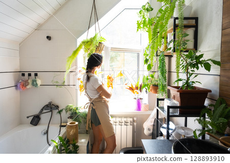 Woman in rubber gloves and apron manually washes window of house in bathroom with rag cleaner and mop inside interior with home plants. Restoring order and cleanliness, cleaning servise 128893910