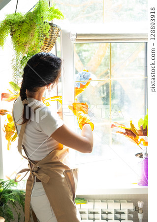 Woman in rubber gloves and apron close-up manually washes window of house with rag cleaner and mop inside interior with home plants on windowsill. Restoring order and cleanliness, cleaning servise 128893918