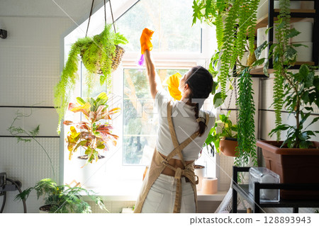 Woman in rubber gloves and apron manually washes window of house in bathroom with rag cleaner and mop inside interior with home plants. Restoring order and cleanliness, cleaning servise Woman in rubber gloves and apron manually washes window of house in bathroom with rag cleaner and mop inside interior with home plants. Restoring order and cleanliness, cleaning servise 128893943