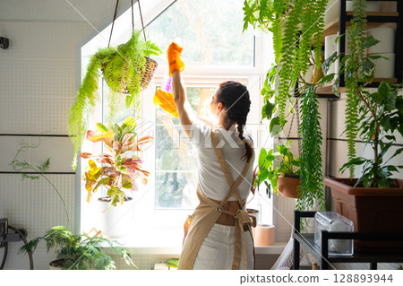Woman in rubber gloves and apron manually washes window of house in bathroom with rag cleaner and mop inside interior with home plants. Restoring order and cleanliness, cleaning servise Woman in rubber gloves and apron manually washes window of house in bathroom with rag cleaner and mop inside interior with home plants. Restoring order and cleanliness, cleaning servise 128893944