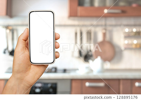 Blurred kitchen background with hand holding phone with empty white blank screen. Smart home control in kitchen by apps in smartphone. Online grocery ordering concept, online food shopping. Space for Blurred kitchen background with hand holding phone with empty white blank screen. Smart home control in kitchen by apps in smartphone. Online grocery ordering concept, online food shopping. Space for 128895176