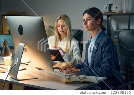 Professional Women Collaborating At Computer In Modern Office Setting At Night 128898130