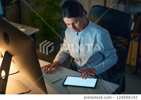 Dedicated Professional Working Late Night Using Tablet and Computer in Office Setting 128898142