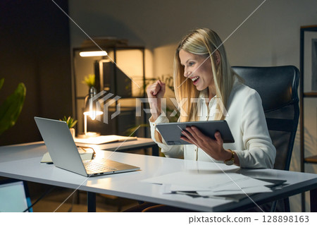 Excited Businesswoman Celebrating Achievement Working Late at Office with Laptop and Tablet 128898163