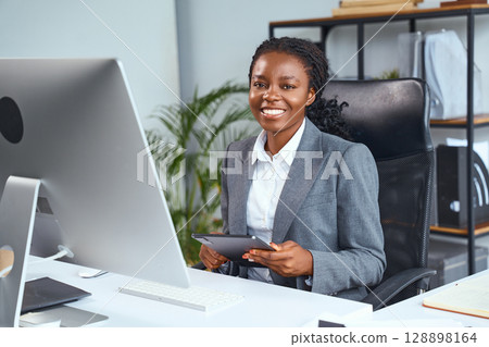 Confident Professional Woman Sitting at Desk Holding Tablet in Office Workspace 128898164