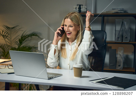 Excited Businesswoman Celebrating Success on Phone Call While Working at Office Desk 128898207