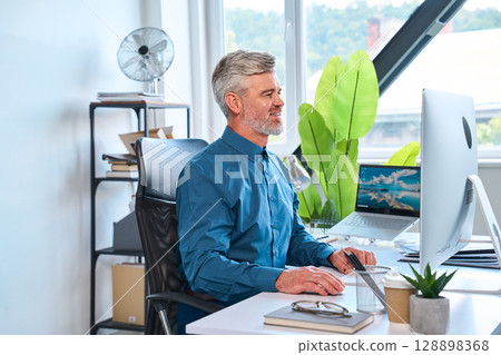 Professional Man Smiling While Working at Modern Office Desk With Computer Monitor 128898368