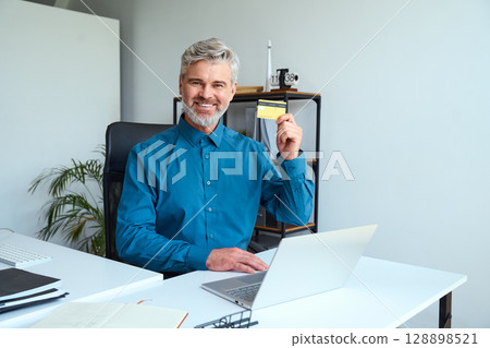 Smiling Man Sitting At Desk Holding Credit Card With Laptop And Notebooks Smiling Man Sitting At Desk Holding Credit Card With Laptop And Notebooks 128898521