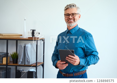 Smiling Professional Holding Tablet in Modern Office Space with Shelves in Background 128898575