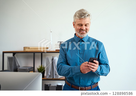 Smiling Mature Businessman Checking His Smartphone in a Modern Office Environment Smiling Mature Businessman Checking His Smartphone in a Modern Office Environment 128898586