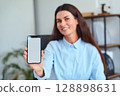 Smiling Woman Displays Smartphone With Blank Screen in a Modern Office Environment 128898631