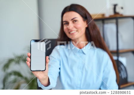 Smiling Woman Displays Smartphone With Blank Screen in a Modern Office Environment Smiling Woman Displays Smartphone With Blank Screen in a Modern Office Environment 128898631