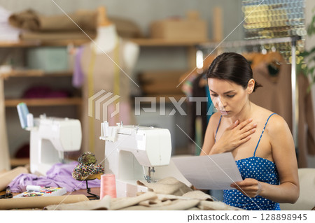 Upset young woman seamstress with documents in workshop Upset young woman seamstress with documents in workshop 128898945