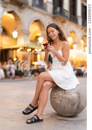 Young woman sitting on stone ball with phone 128899023