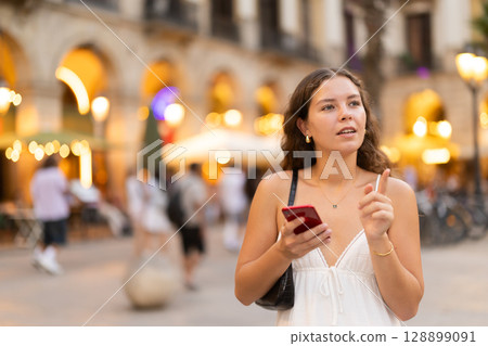 In evening, smiling girl with smartphone walks along city street Placa Real In evening, smiling girl with smartphone walks along city street Placa Real 128899091