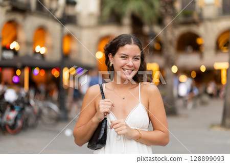 In evening, young smiling girl walks along city street Placa Real 128899093