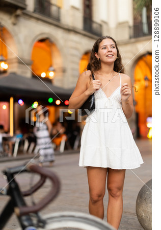 In evening, young smiling girl walks along city street Placa Real 128899106