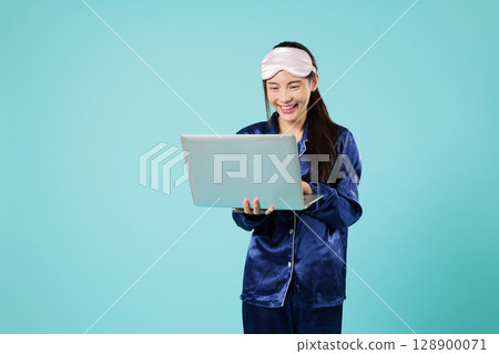 Happy woman in pajama sleep mask remote work Asian smile with laptop chatting and working from home, relax good mood using the laptop pc computer, headshots isolated studio blue background 128900071