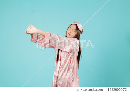 Asian woman stretching arms up in pajamas, sleep mask on head, a joyful early morning awakening after a pleasant sleep, eyes closed, enjoys a good day, headshots isolated studio blue background 128900072