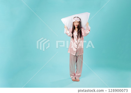 Full body young asian woman in pajamas smiles, holding pillow behind her head to help her sleep as she dreams of a good mood night nap and a restful sleep, isolated studio blue background Full body young asian woman in pajamas smiles, holding pillow behind her head to help her sleep as she dreams of a good mood night nap and a restful sleep, isolated studio blue background 128900073
