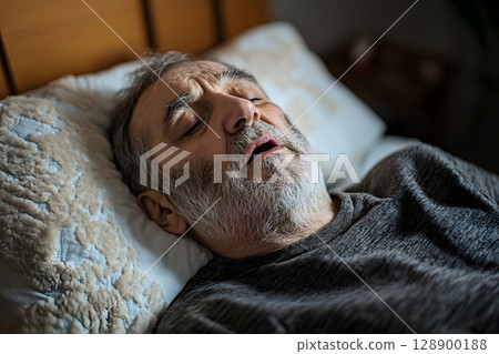 A mature man sleeps heavily in his comfortable bed with an open mouth, likely snoring during the night. 128900188