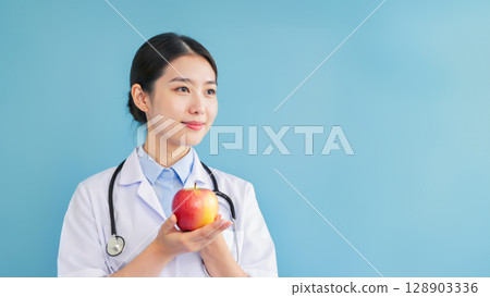 Smiling female doctor in white coat holding red apple against blue background. Concept of healthy lifestyle, preventive care, nutrition awareness, and wellness education in modern healthcare Smiling female doctor in white coat holding red apple against blue background. Concept of healthy lifestyle, preventive care, nutrition awareness, and wellness education in modern healthcare 128903336