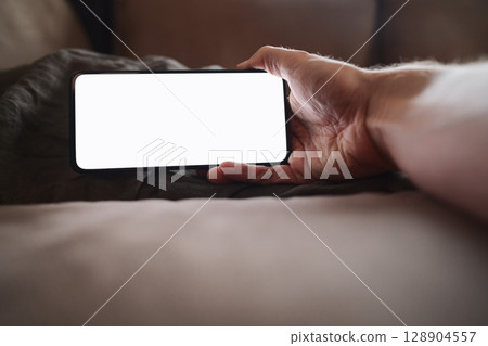 man hand holding phone with white screen over bed on background man hand holding phone with white screen over bed on background 128904557