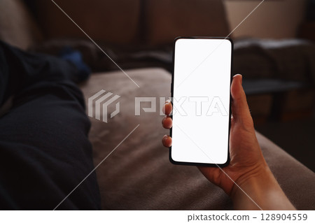 Man hold smartphone with white screen in his hand lying on a couch in home environment Man hold smartphone with white screen in his hand lying on a couch in home environment 128904559